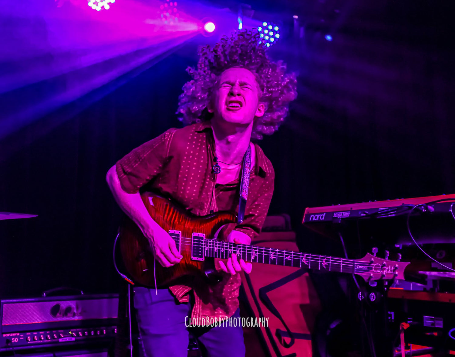 Isaac Hadden making a solid stank face as he shreds his guitar on a colorfully lit indoor stage.