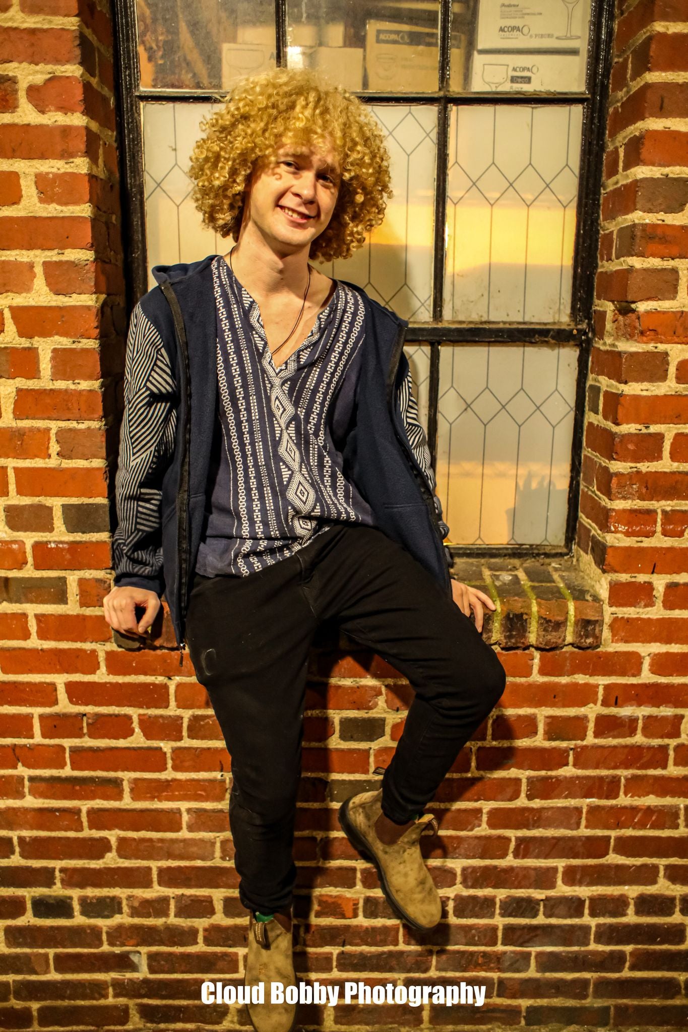 Isaac Hadden sitting on a brick wall smiling at the camera, a young man with curly blonde hair wearing a trippy T-shirt.