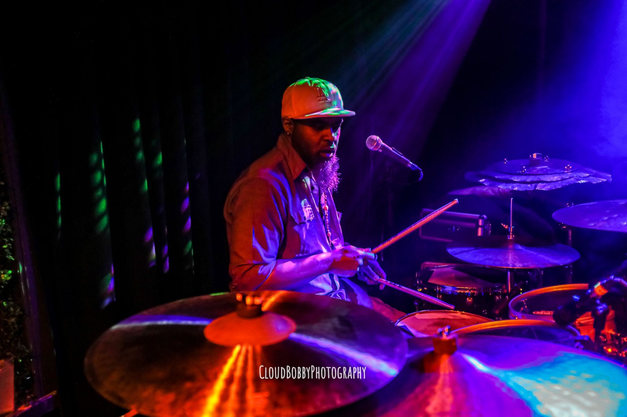 Iajhi Hampden making a solid stank face as he shreds his drums on a colorfully lit indoor stage. 