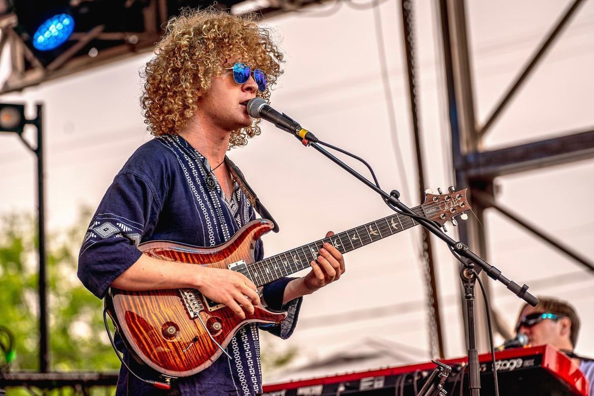 Isaac Hadden wearing a funky fresh colorful T-shirt while playing a solo on guitar on an outdoor stage at a festival. 