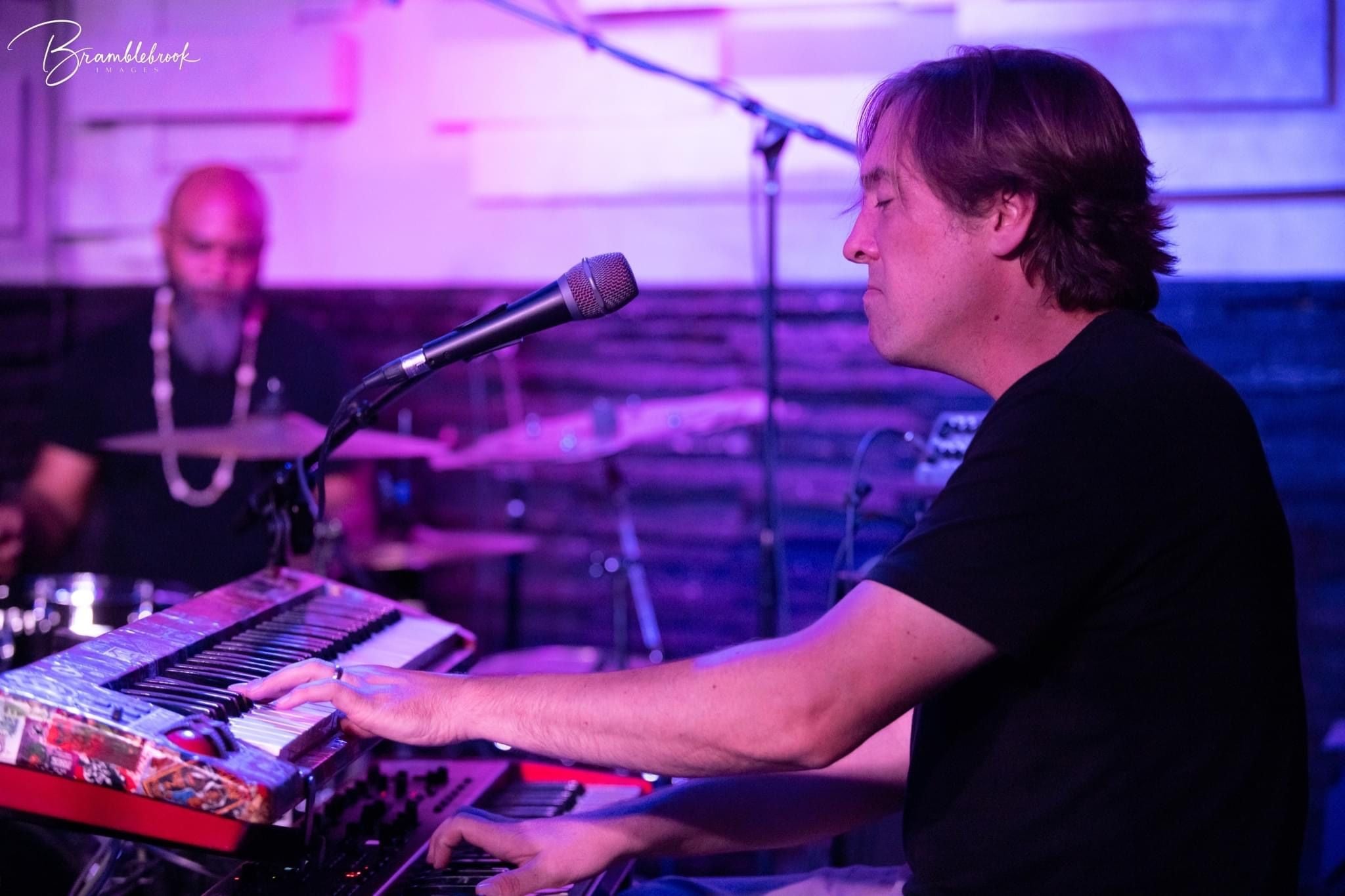 Bill Stevens playing the keys on  an colorfully lit indoor stage, wearing an Isaac Hadden Organ Trio black T-shirt..